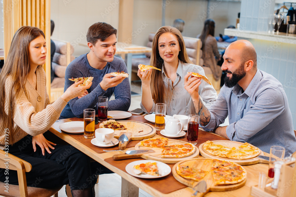 A group of young cheerful friends is sitting in a cafe talking and eating pizza. Lunch at the pizzeria. A group of young cheerful friends is sitting in a cafe talking and eating pizza. Lunch at the pizzeria.