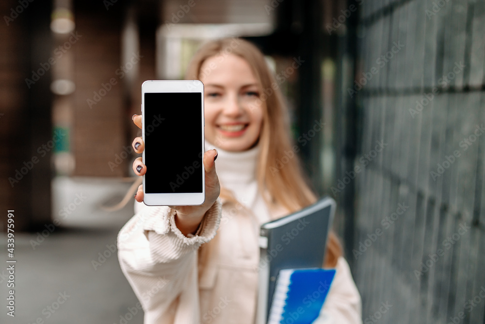 Happy smiling blonde student girl showing screen of a mobile phone against the background of the college. Mockup for design