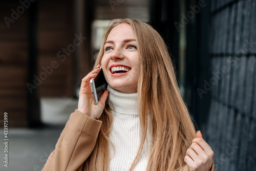 Young blonde Caucasian woman in a beige coat talking on a mobile phone  smiling and looking up against the background of an office building. Corporate employee. Happy female IT-specialist