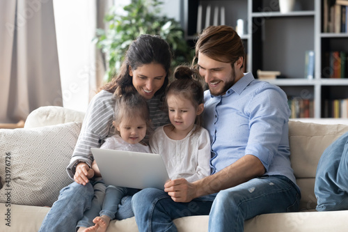 Smiling young Caucasian family with two little daughters relax on couch at home use laptop. Happy parents and small girls children have fun talk on webcam video call on computer. Technology concept.