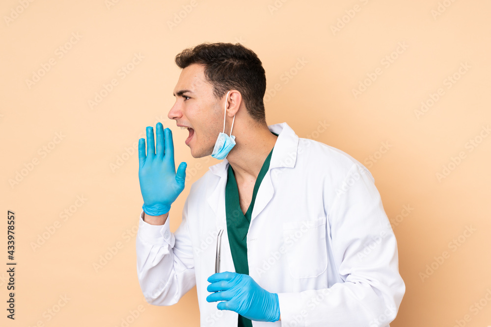 Young dentist man holding tools isolated on beige background shouting with mouth wide open to the lateral Young dentist man holding tools isolated on beige background shouting with mouth wide open to the lateral