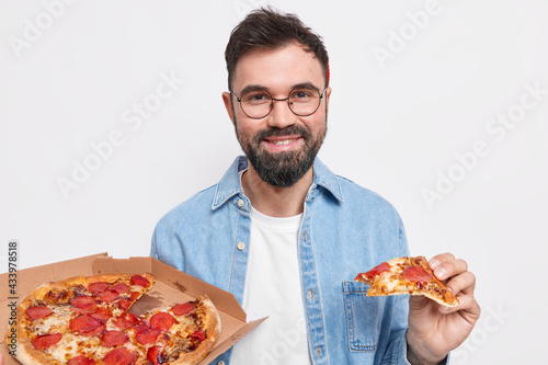 Pleased handsome bearded man eats delicious pizza for dinner feels hungry wears round glasses and shirt eats junk food isolated over white background. Cheat meal concept. Having delicious dinner