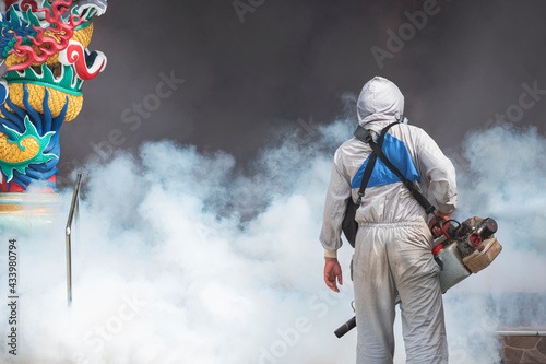 Rear view of outdoor healthcare worker in protective clothing using fogging machine spraying chemical to eliminate mosquitoes and prevent dengue fever at chinese shrine in Samutsakhon, Thailand