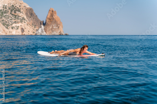 Sporty girl on a glanders surfboard in the sea on a sunny summer day. In a striped swimsuit, lying on his stomach, rowing with his hands. Summer activities by Stortom by the sea.