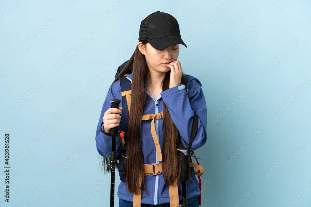 Young Chinese girl with backpack and trekking poles over isolated blue background having doubts Young Chinese girl with backpack and trekking poles over isolated blue background having doubts