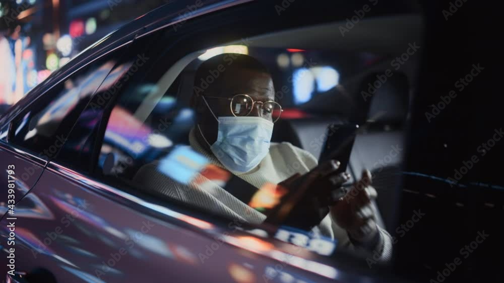 Stylish Black Man Wearing a Face Mask Commuting Home in a Backseat of a Taxi at Night. Handsome Male Making a Video Call on a Smartphone while in Transfer Car on City Street with Working Neon Signs.
