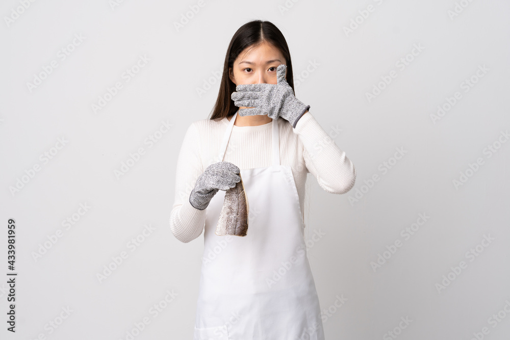 Chinese fishmonger wearing an apron and holding a raw fish over isolated white background covering mouth with hand Chinese fishmonger wearing an apron and holding a raw fish over isolated white background covering mouth with hand