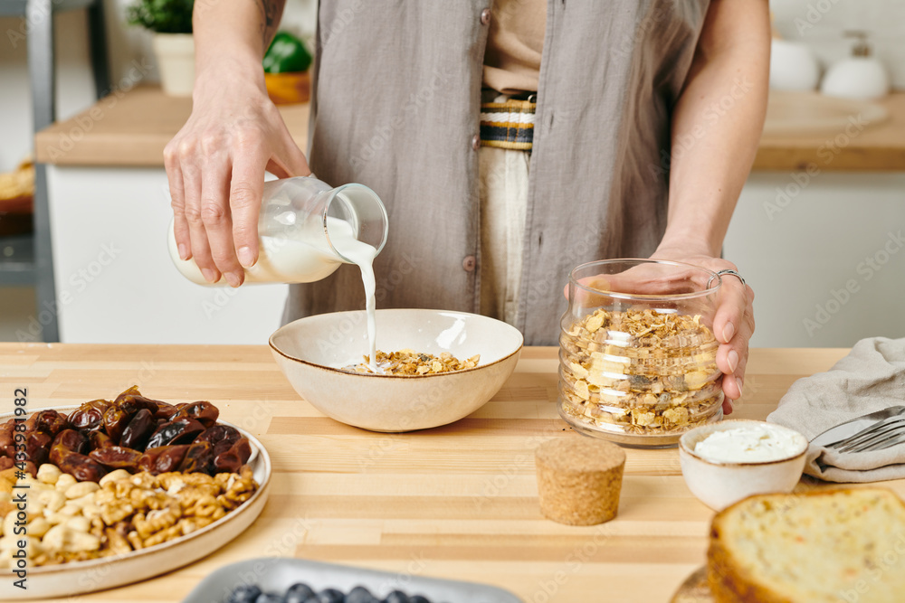 A woman pouring milk into bowl with corn flakes by kitchen table