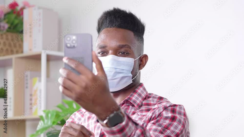 Coronavirus. African man in quarantine for coronavirus wearing protective mask, He is working from home and using mobile phone at sofa. Video call. Video conference with mobile phone Coronavirus. African man in quarantine for coronavirus wearing protective mask, He is working from home and using mobile phone at sofa. Video call. Video conference with mobile phone