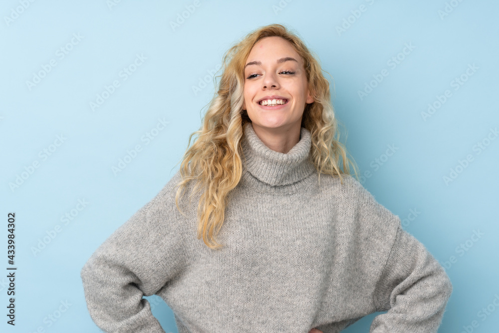 Young blonde woman wearing a sweater isolated on blue background posing with arms at hip and smiling Young blonde woman wearing a sweater isolated on blue background posing with arms at hip and smiling