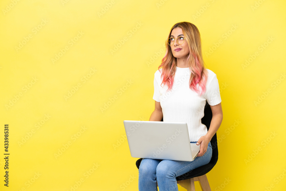 Naklejka premium Young woman sitting on a chair with laptop over isolated yellow background and looking up