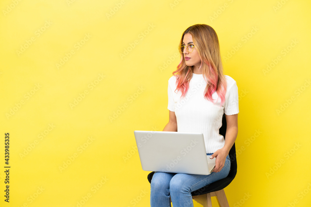 Young woman sitting on a chair with laptop over isolated yellow background looking to the side Young woman sitting on a chair with laptop over isolated yellow background looking to the side