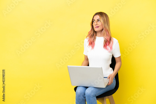 Young woman sitting on a chair with laptop over isolated yellow background and looking up © luismolinero