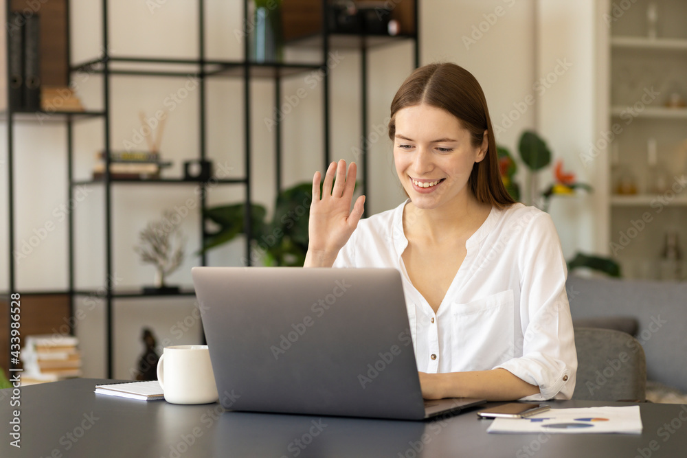 Smiling young Caucasian woman sitting at her office desk wave greet talk on video call online . Zoom conference concep Smiling young Caucasian woman sitting at her office desk wave greet talk on video call online . Zoom conference concep