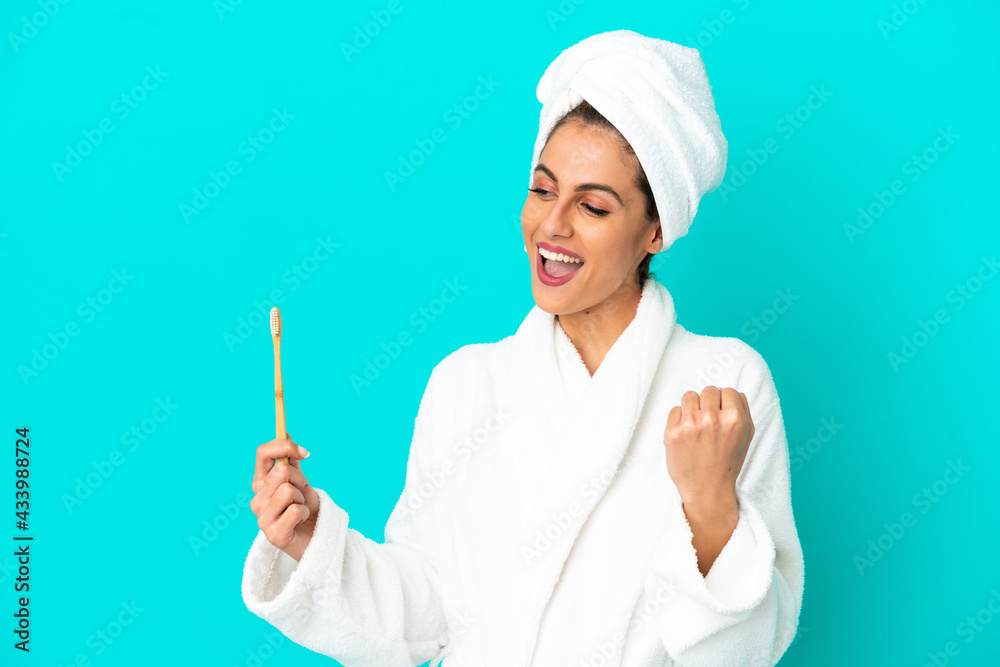 Young woman in a bathrobe brushing teeth over isolated blue background celebrating a victory Young woman in a bathrobe brushing teeth over isolated blue background celebrating a victory