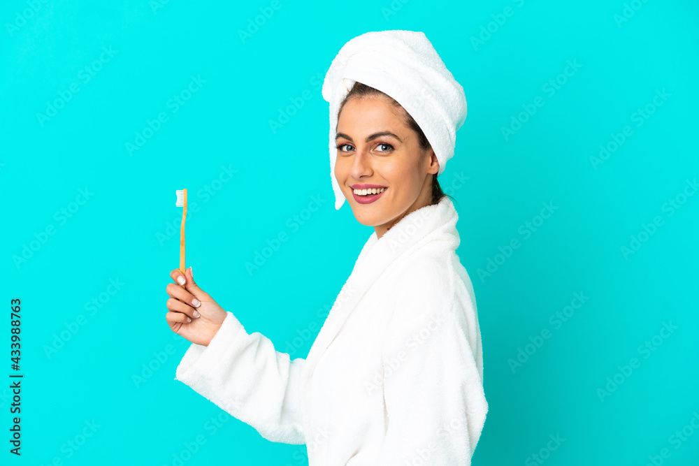 Young woman in a bathrobe brushing teeth over isolated blue background smiling a lot Young woman in a bathrobe brushing teeth over isolated blue background smiling a lot