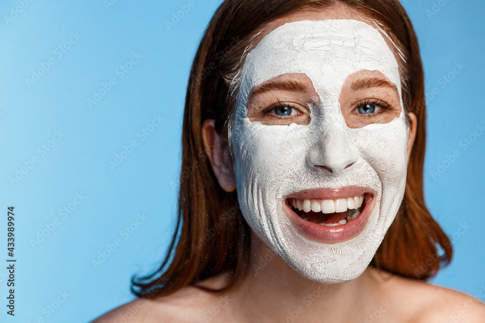 Young girl with facial mask looking at camera over white background. Cosmetic procedure. Beauty spa and cosmetology. Redhead woman laughing and smiling during skincare facial procedure Young girl with facial mask looking at camera over white background. Cosmetic procedure. Beauty spa and cosmetology. Redhead woman laughing and smiling during skincare facial procedure