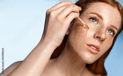 Woman applying hyaluronic serum on her face with pipette. Photo of attractive woman with perfect makeup blue background. Beauty concept. Redhead girl with freckles drop essential oil on facial skin photo