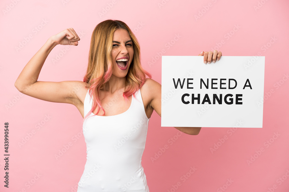 Young woman over isolated pink background holding a placard with text We Need a Change and doing strong gesture Young woman over isolated pink background holding a placard with text We Need a Change and doing strong gesture