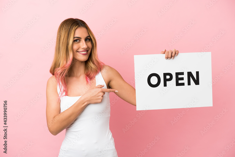 Young woman over isolated pink background holding a placard with text OPEN and pointing it Young woman over isolated pink background holding a placard with text OPEN and pointing it