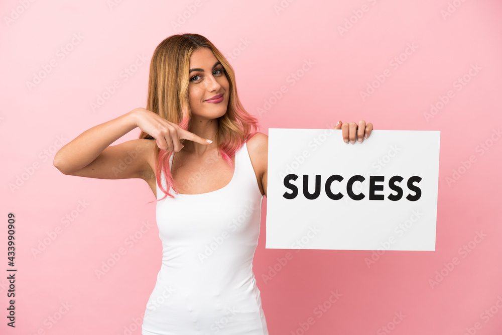 Young woman over isolated pink background holding a placard with text SUCCESS and pointing it Young woman over isolated pink background holding a placard with text SUCCESS and pointing it