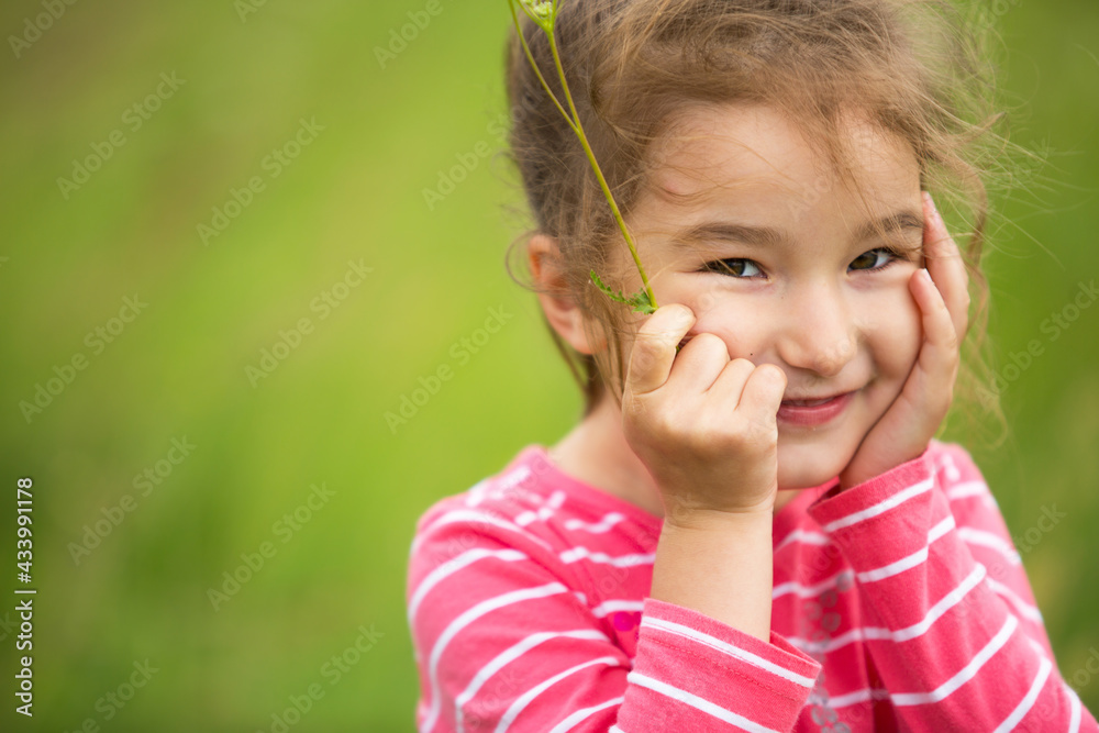 Little girl in a coral striped T-shirt on a green background in a field holds her face in her hands and smiles slyly. Children's Day, happy child, environmental and nature protection, insect repellent Little girl in a coral striped T-shirt on a green background in a field holds her face in her hands and smiles slyly. Children's Day, happy child, environmental and nature protection, insect repellent