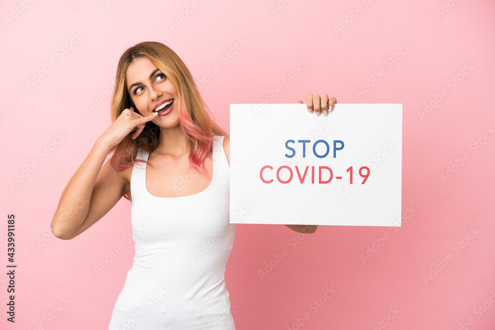 Young woman over isolated pink background holding a placard with text Stop Covid 19 and doing phone gesture Young woman over isolated pink background holding a placard with text Stop Covid 19 and doing phone gesture