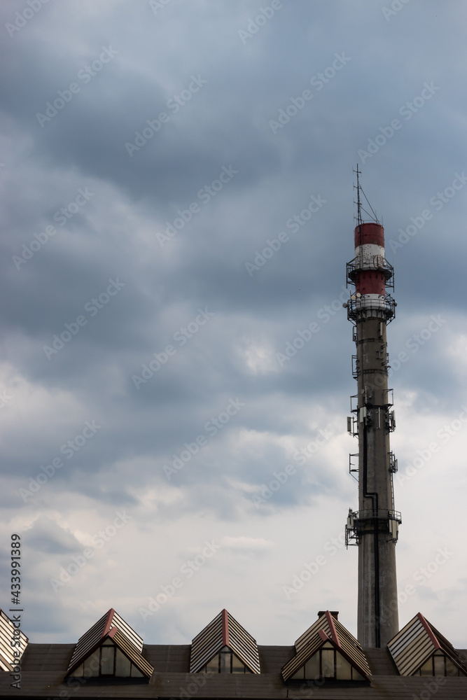 Rooftop of industrial halls and an inactive municipal heating plant chimney converted into a mast for telecommunications antennas against the cloudy sky. The photo was taken in natural daylight.