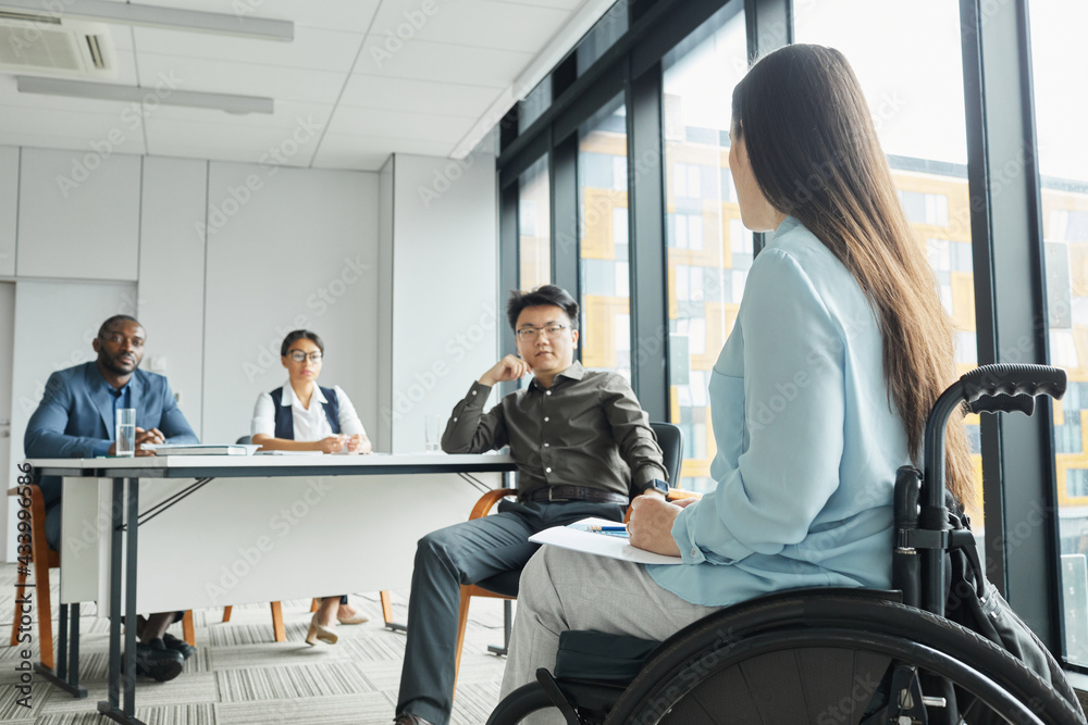 Side view portrait of young woman in wheelchair giving speech to diverse business team in office, copy space Side view portrait of young woman in wheelchair giving speech to diverse business team in office, copy space