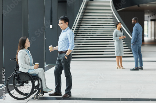 Full length side view portrait of young businesswoman in wheelchair chatting with male colleague in office lobby, copy space