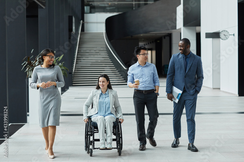 Full length view at diverse group of business people chatting in graphic office lobby while moving towards camera, focus on smiling young woman in wheelchair photo