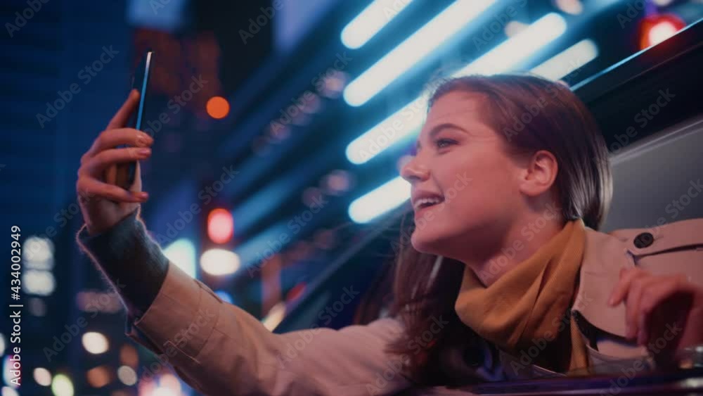 Excited Female is Looking Out of the Window from Backseat of a Car at Night. Woman Taking Photos and Videos on a Smartphone with Amazement of How Beautiful is the City Street with Working Neon Signs. Excited Female is Looking Out of the Window from Backseat of a Car at Night. Woman Taking Photos and Videos on a Smartphone with Amazement of How Beautiful is the City Street with Working Neon Signs.