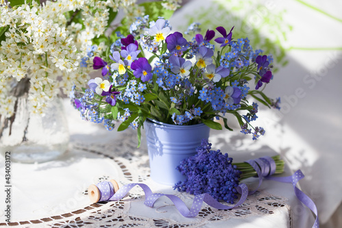 A bouquet of spring blue forget-me-not flowers and pansies in a vase on the table, tablecloth, muscari, bird cherry and ribbon.