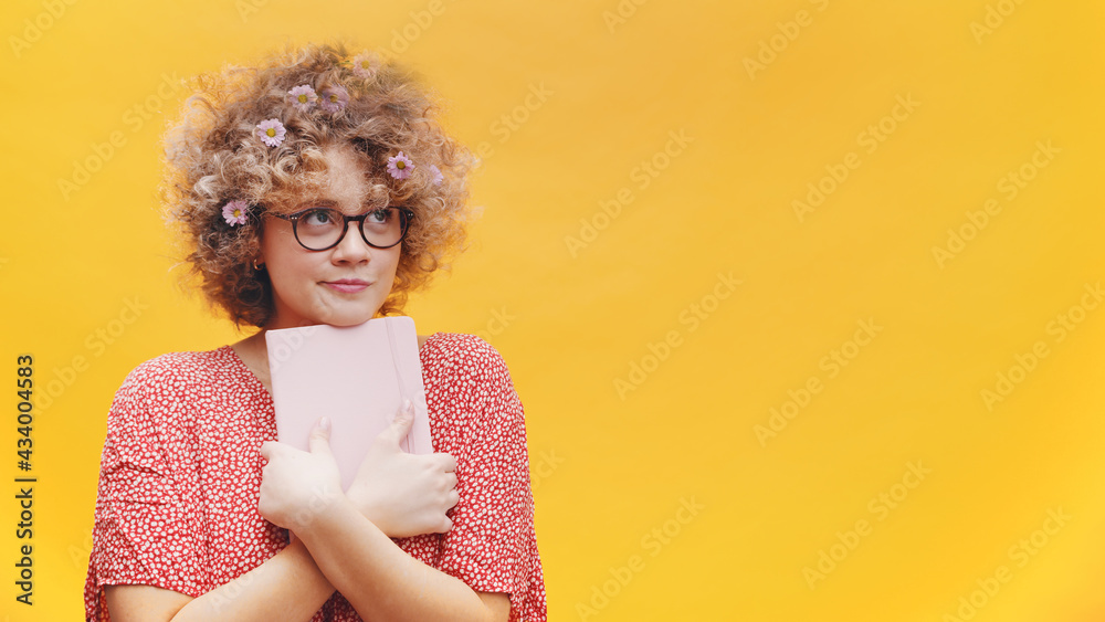 Shy and excited young woman with flowers in the hair hugging her book and making funny faces. Isolated on yellow background. Copy space. High quality photo Shy and excited young woman with flowers in the hair hugging her book and making funny faces. Isolated on yellow background. Copy space. High quality photo
