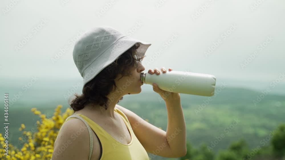 Female hiker with hat drink water from metal bottle during outdoors hike. Woman drinks water during hot summer day in nature making sure not to dehydrate while traveling outside. Green hills in BG 