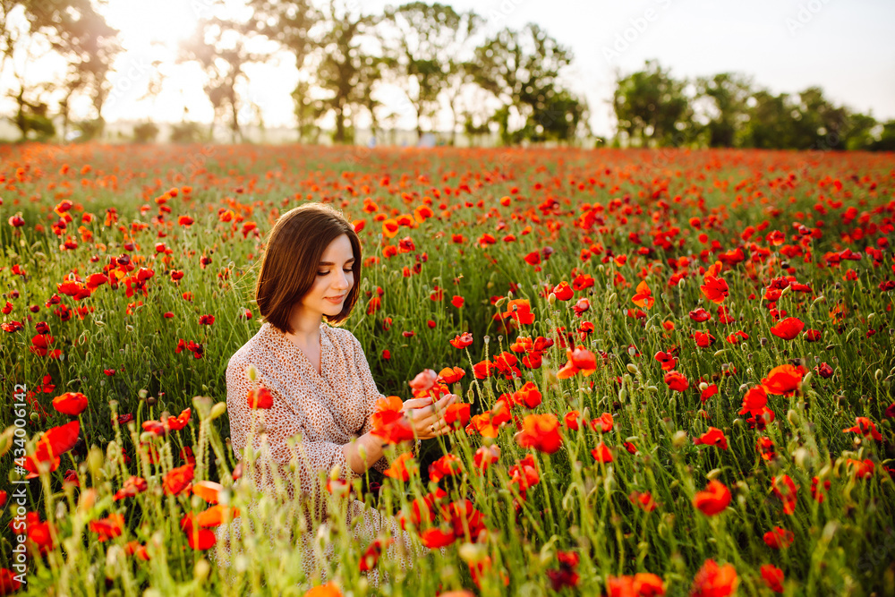 Fototapeta premium Beautiful young woman enjoys sitting among red poppy flowers on the field on the sunset. A girl feels harmony with nature and touches the plants. Love, peace and satisfaction concept.