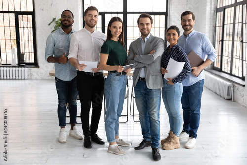 Portrait of happy millennial diverse professional team in loft office space. Group of multi ethnic employees gathering for corporate meeting and teamwork, looking at camera, smiling. Full length photo