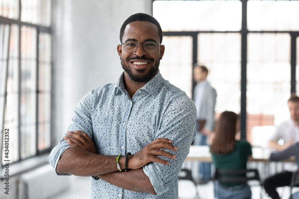 Portrait of happy African American small business owner posing with hands folded. Millennial black male team leader smiling, looking at camera, employees working in modern office behind. Head shot Portrait of happy African American small business owner posing with hands folded. Millennial black male team leader smiling, looking at camera, employees working in modern office behind. Head shot
