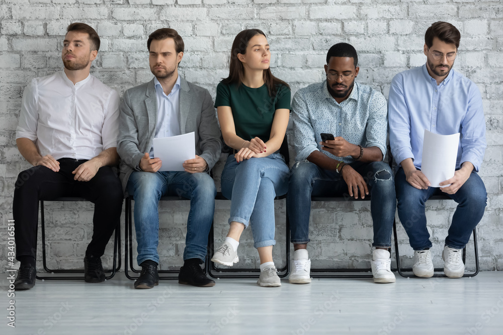 Fototapeta premium Bored vacancy applicant waiting for appointment with employee too long, sitting on chairs in corridor, holding paper resume, using smartphone, looking away. Candidates expecting job interview