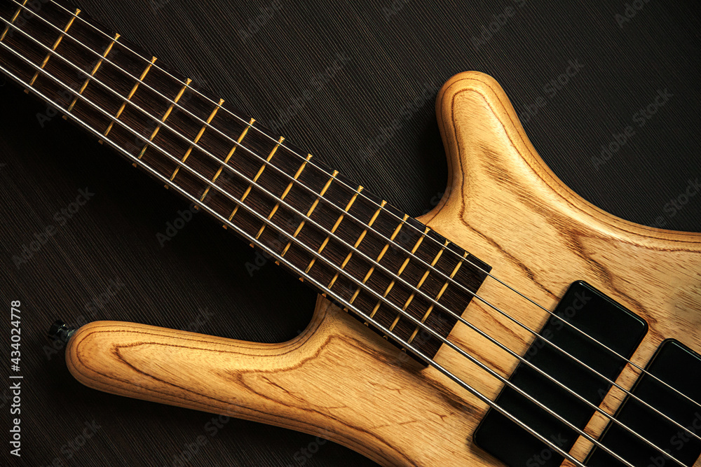 Fototapeta premium Close up of electric bass guitar with five strings. Detailed view of wooden varnished texture of musical instrument on dark brown background. Music classes, concert, hobby and bass player concept 
