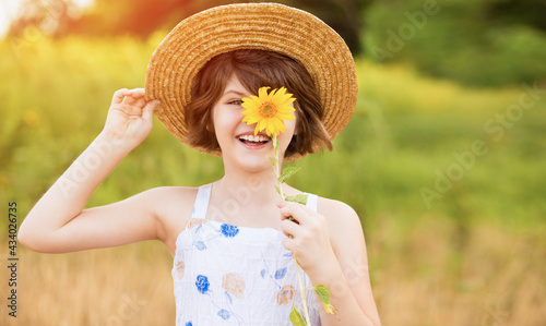 Beautiful little girl in straw hat with fluttering hair smile and hide eye with sunflower flower, walking outdoor in summer holiday