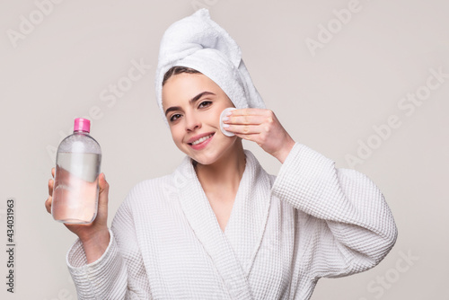 Happy young woman in bathrobe with towel on head cleansing face with micellar water and cotton pad isolated on pink. Girl with micellar water. photo
