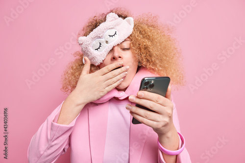 Tired woman with curly hair feels exhausted after having corporate meeting very late checks information in smartphone covers mouth yawns wears sleepmask travel pillow around neck fomal clothes photo