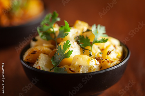 Close-up of Indian vegetarian classic dish Jeera Aloo - Potatoes Flavored with Cumin garnished with green coriander fresh leaves. Served in black ceramic bowl over wooden background