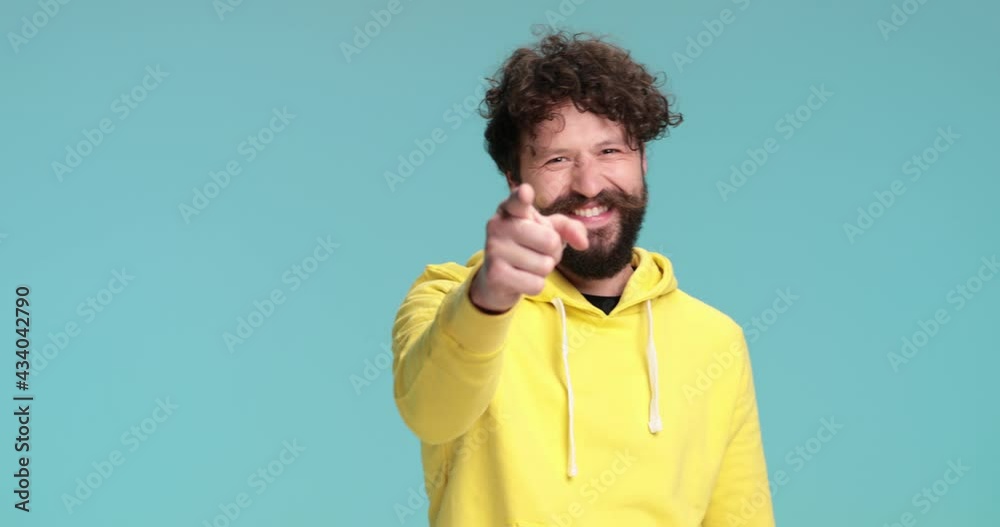 casual young man with long beard and curly hair wearing yellow hoodie, pointing fingers and smiling on blue background in studio casual young man with long beard and curly hair wearing yellow hoodie, pointing fingers and smiling on blue background in studio