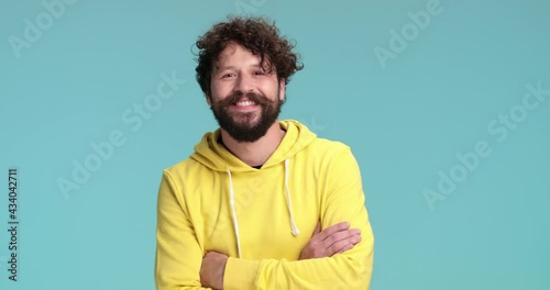 cool long bearded guy in yellow hoodie smiling, nodding and crossing arms while standing on blue background in studio photo