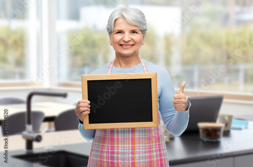 cooking  culinary and old people concept - portrait of smiling senior woman in apron holding chalkboard and showing thumbs up over home kitchen on background