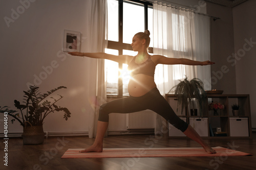 sport, fitness and people concept - happy pregnant woman doing yoga warrior pose at home over sunshine