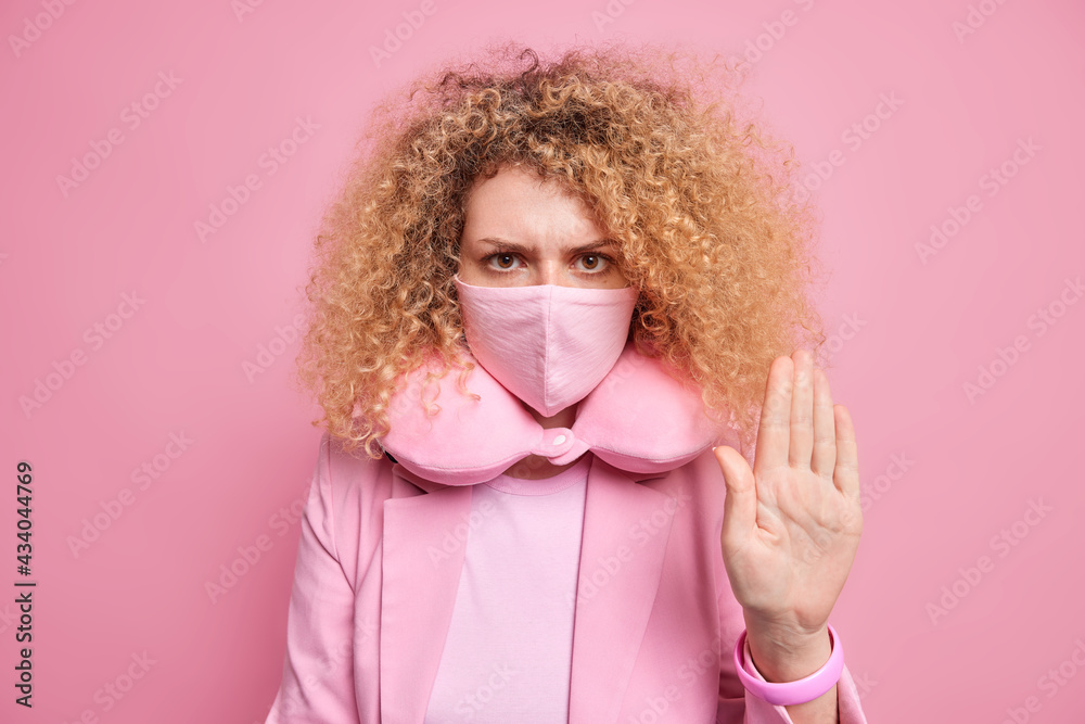 Serious curly haired woman asks not to forget about mask makes stop gesture demands to keep social distance uses safety protective items during coronavirus pandemic outbreak wears neck pillow Serious curly haired woman asks not to forget about mask makes stop gesture demands to keep social distance uses safety protective items during coronavirus pandemic outbreak wears neck pillow