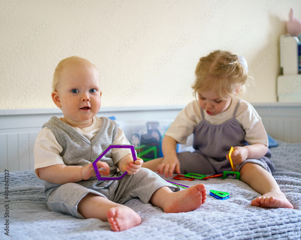 Cute little children infant boy and toddler girl having fun while playing with toy construction set on bed at home. Playtime for curious brother and sister in bedroom. Family spending time together Cute little children infant boy and toddler girl having fun while playing with toy construction set on bed at home. Playtime for curious brother and sister in bedroom. Family spending time together